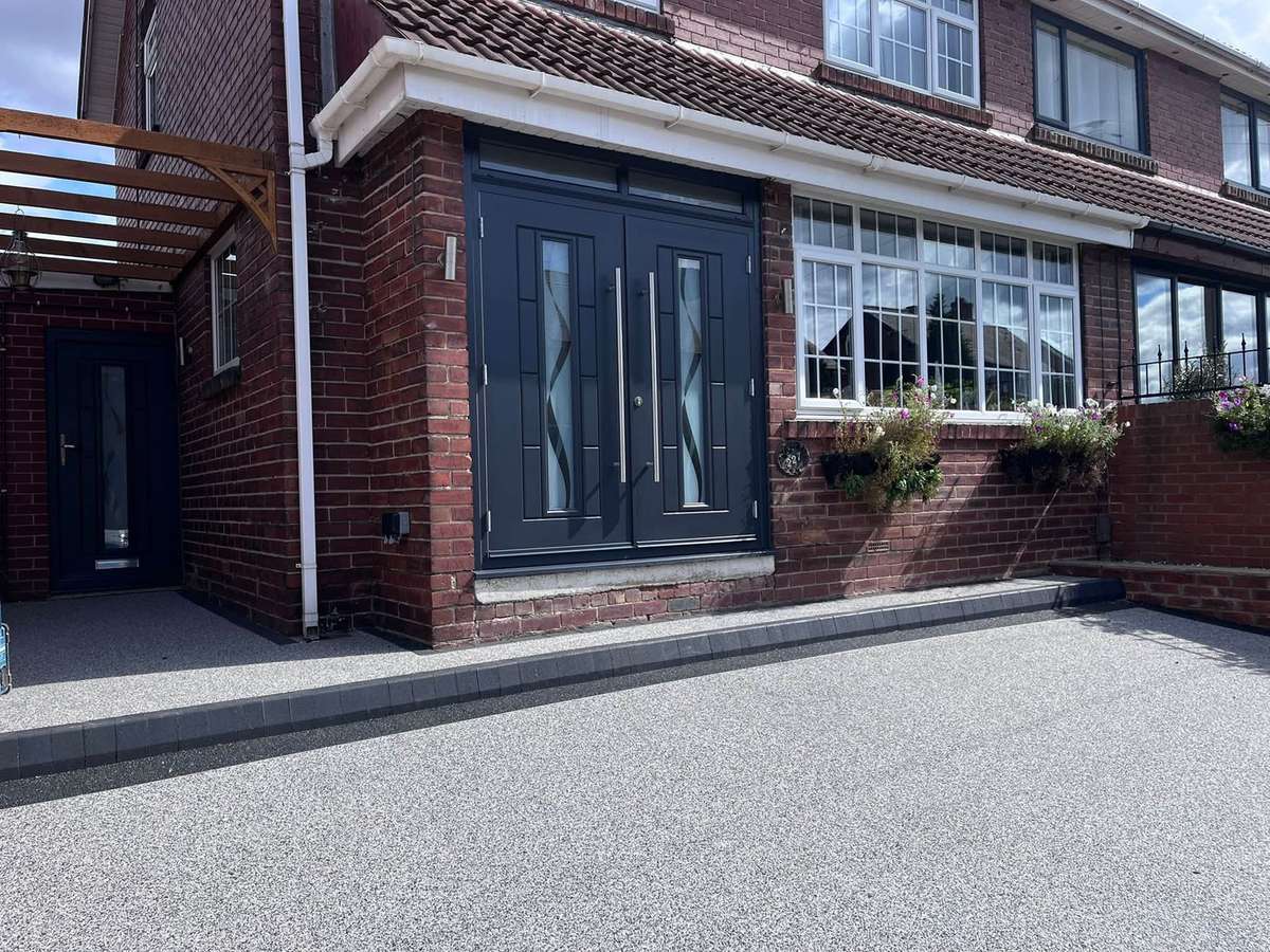 An anthracite composite double door with brushed bar handles beside a white bay window, on a red brick house with a new resin driveway